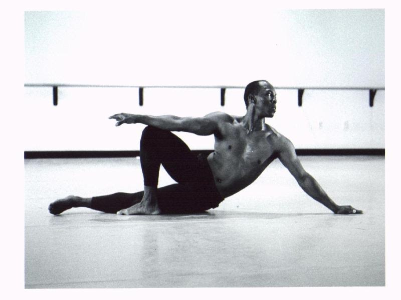 Black-and-white photograph of Boris Willis seated on the studio floor, one arm extended, beside a ballet barre